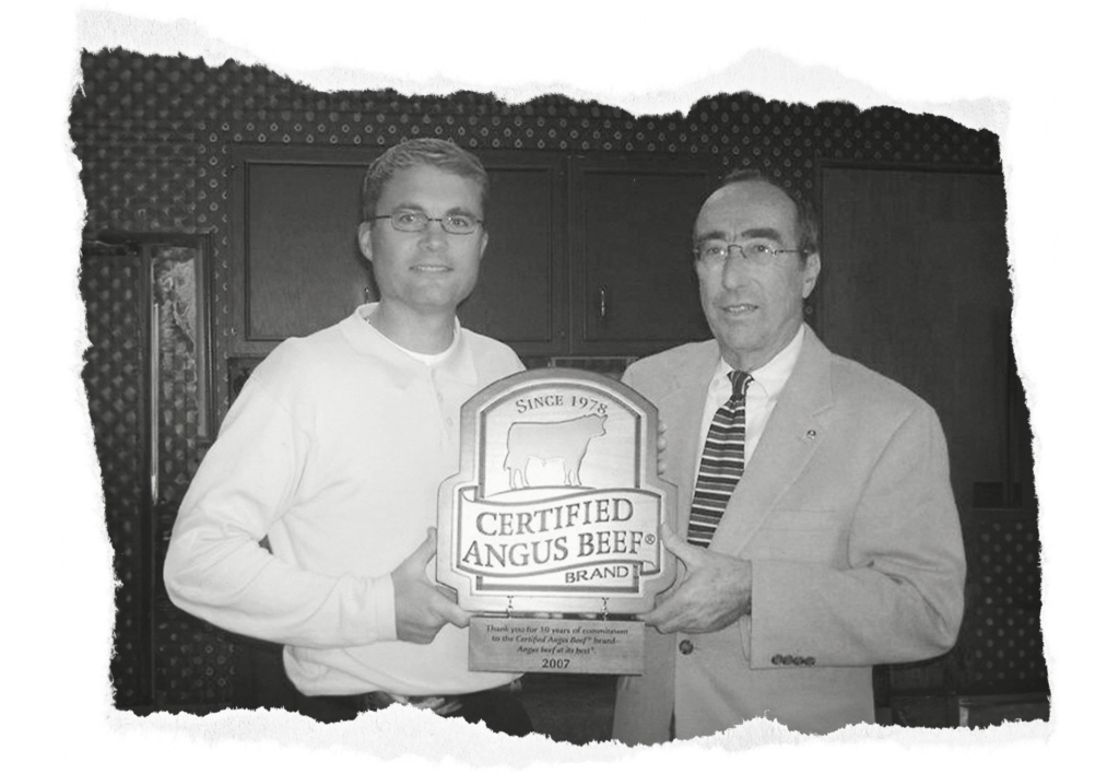 Two men are holding a "Certified Angus Beef" plaque together. The person on the left is wearing a light-colored long-sleeve shirt and glasses, while the man on the right is in a suit and tie. The background includes a wall with a patterned design. The image is in black and white.