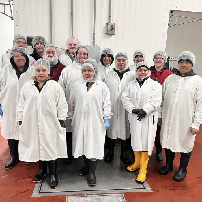 A group of 14 people, wearing white coats, hairnets, and black boots (one person has yellow boots), stands on a red floor inside the WG-Provisions facility in Iowa. Most have their hands behind their backs or in front of them as they pose for the photo against the sterile, white-walled background.