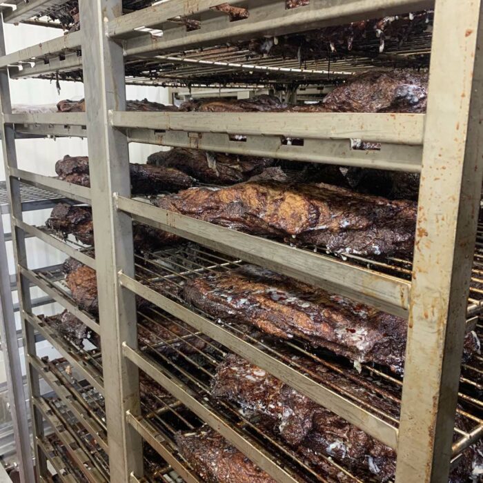 Multiple metallic racks filled with smoked meat, arranged in a vertical industrial-style setup. The meat appears dark and glossy, with a distinct smoky texture indicative of a slow and thorough cooking process. The background shows the clean walls of WG-Provisions' Iowa kitchen or processing area.