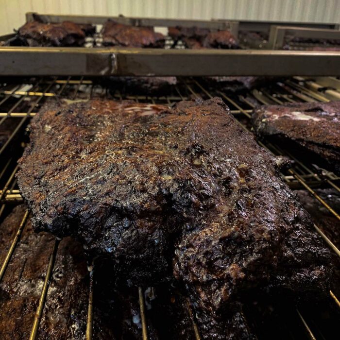 Close-up of several large, dark brown cuts of brisket, heavily charred and smoked, resting on metal racks inside a WG-Provisions commercial smoker in Iowa. The meat showcases a well-developed crust from the smoking process.
