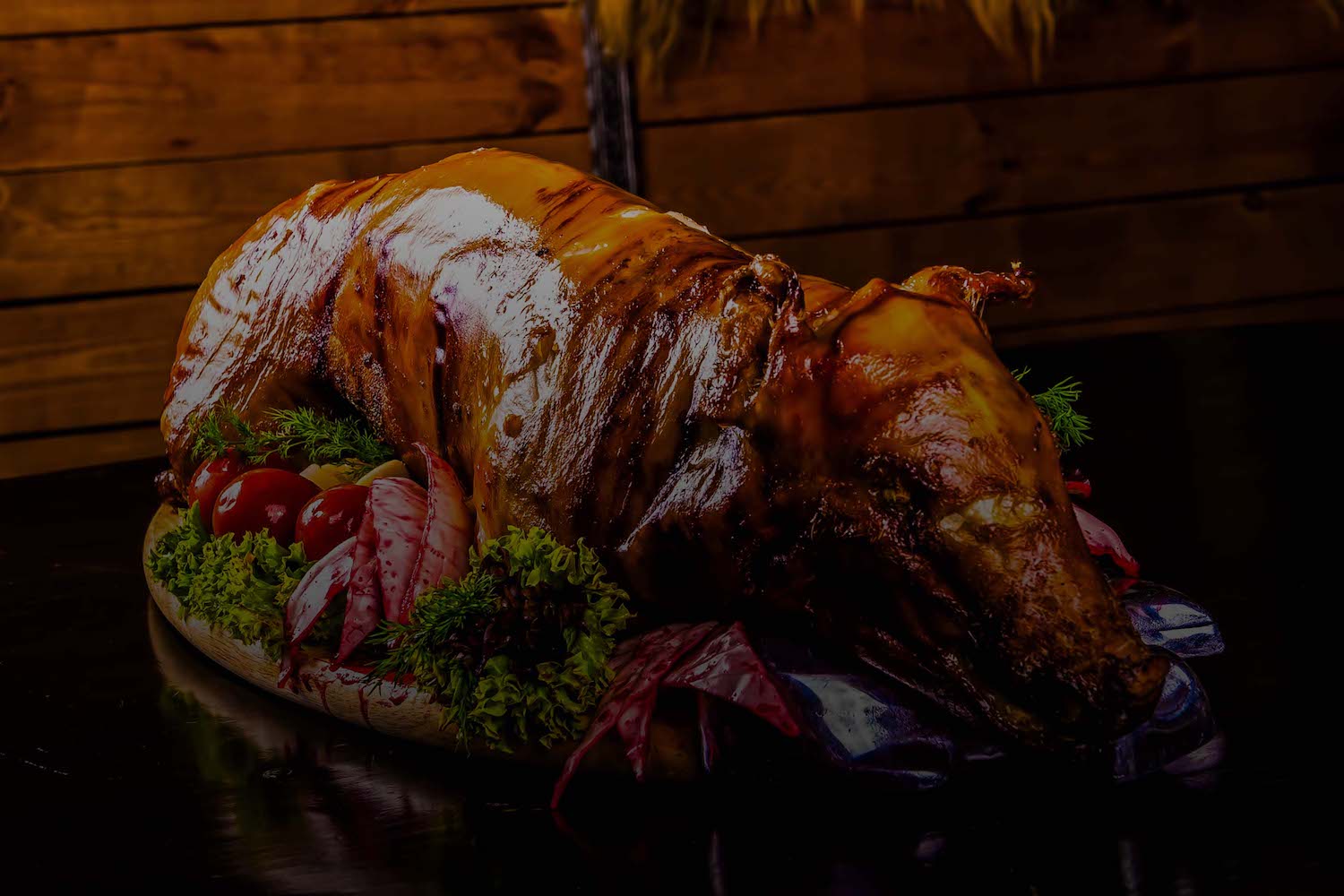 A roasted whole pig with a shiny, glazed exterior is placed on a platter, surrounded by decorative greens, red leaves, and cherry tomatoes. The background features wooden paneling, indicating a warm, rustic setting.