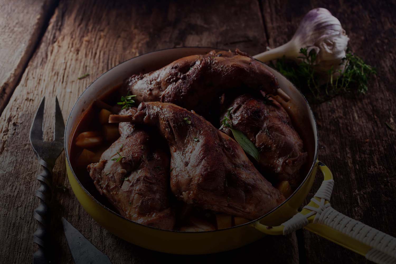 A rustic dish features roasted meat seasoned with herbs, served in a yellow-handled pan on a wooden surface. A garlic bulb and sprigs of fresh thyme lie beside the pan, while cutlery with saw-like edges is positioned on the left. The lighting is dim and moody.