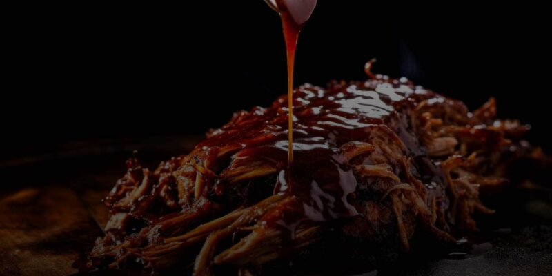 Image of succulent pulled pork on a wooden board, being drizzled with rich, dark barbecue sauce from a metal container by WG-Provisions in Iowa. The glistening sauce highlights the tender and juicy texture of the shredded meat, set against a dark background.