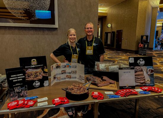Two smiling people stand behind a display table covered with various meat products from Iowa, brochures, and promotional items. The backdrop features a large, modern wall decoration and a partial view of a hotel lobby area. The well-lit and inviting table proudly showcases WG-Provisions' offerings.