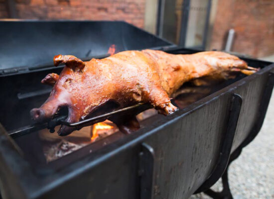 A whole pig is being roasted on a large rotisserie grill outdoors in Iowa. The pig is skewered on a spit, its skin crispy and golden brown with a fire beneath it. The WG-Provisions grill stands on a gravel surface in front of a brick wall, promising a feast to remember.