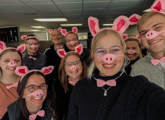 A group of people pose for a photo wearing pig snouts, pink bow ties, and pig ear headbands. They are all smiling and standing close together in an indoor setting with ceiling lights and office decor in the background, showcasing their fun side during the WG-Provisions event in Iowa.