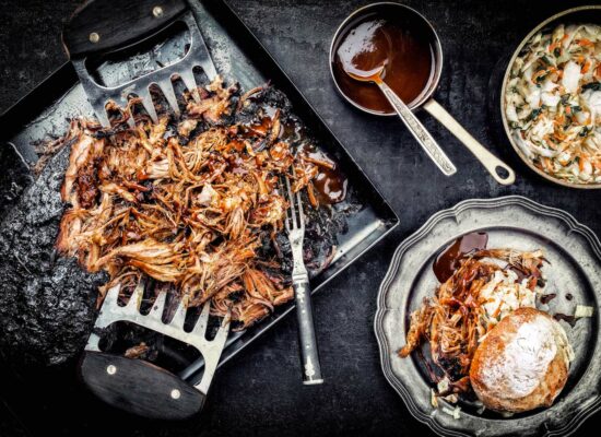 A tray of shredded pulled pork with WG-Provisions metal claws, a fork, and a small pot of barbecue sauce next to it. On the side sits Iowa-style coleslaw and a plate with a pulled pork sandwich topped with coleslaw and sauce. The background is dark and rustic.