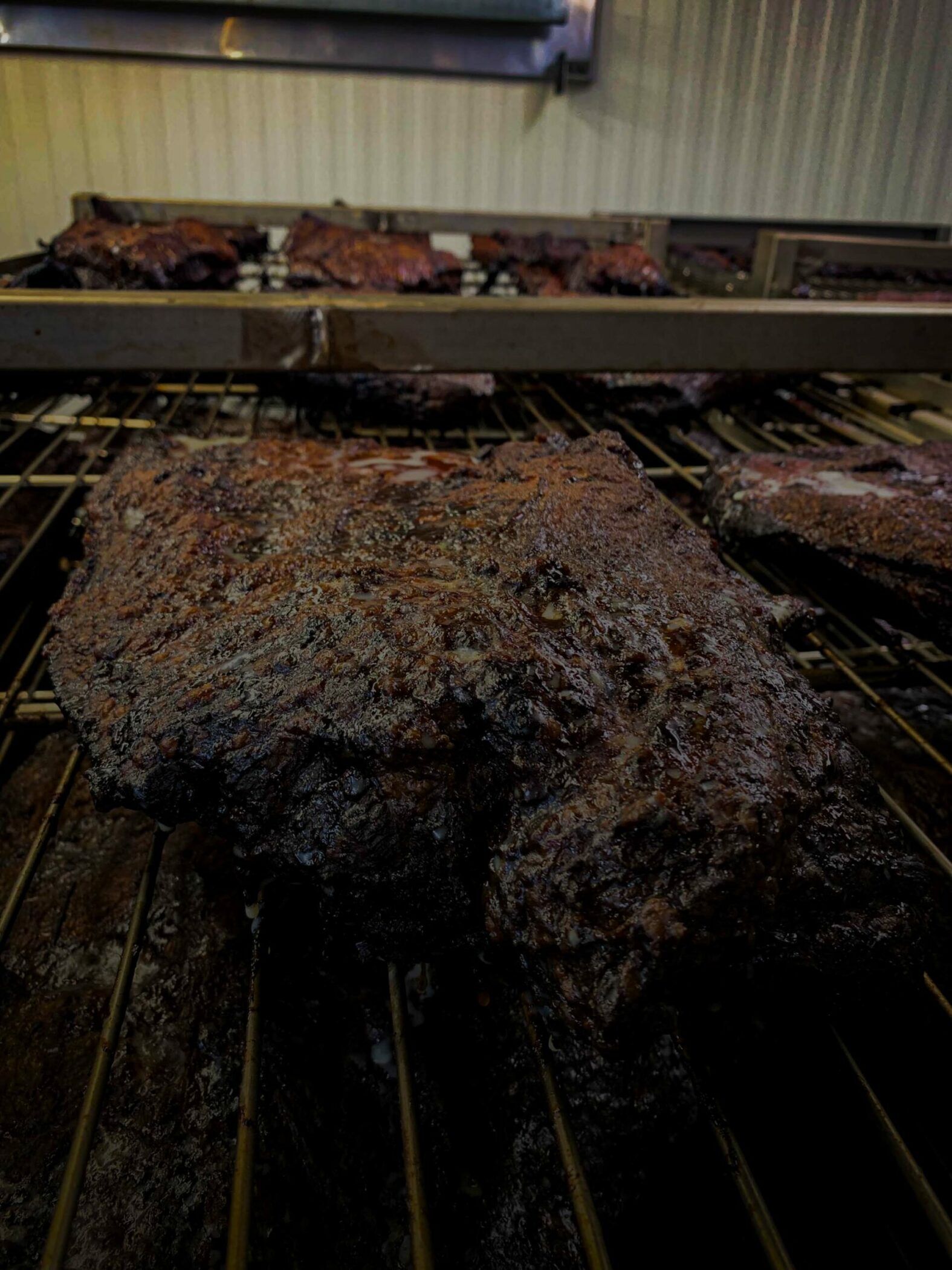 Close-up image of a large piece of meat being smoked on a grill with more pieces cooking in the background. The meat has a dark, crispy exterior, indicating it has been well-cooked and smoked to perfection. This delicious scene unfolds at an Iowa indoor barbecue pit, known for its WG-Provisions quality meats.