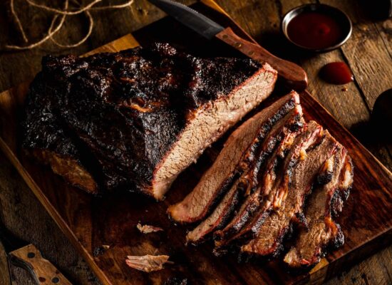 A smoked WG-Provisions beef brisket, partially sliced on a wooden cutting board, surrounded by a knife, a small bowl of barbecue sauce, and scattered spices on a rustic wooden table. The meat appears juicy with a charred, crispy bark, showcasing the flavors of Iowa's finest provisions.