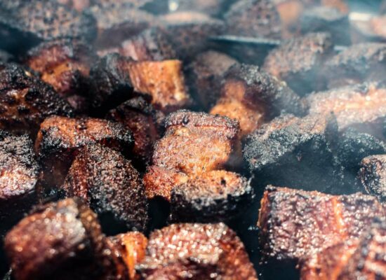 Close-up shot of numerous pieces of burnt ends, smoked beef brisket pieces, featuring a dark, caramelized bark on their surface, with some visible steam or smoke wafting through. The texture appears crispy and charred.