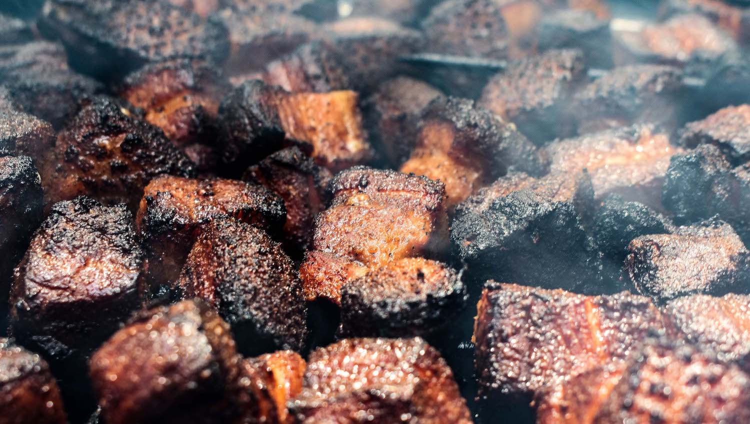 Close-up shot of numerous pieces of burnt ends, smoked beef brisket pieces, featuring a dark, caramelized bark on their surface, with some visible steam or smoke wafting through. The texture appears crispy and charred.