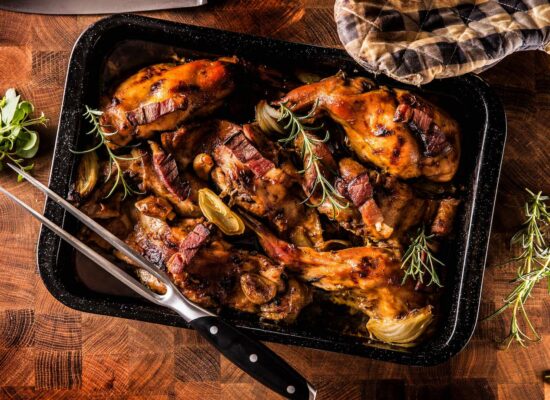 A roasting pan filled with WG-Provisions' cooked rabbit meat topped with bacon strips, caramelized onions, and rosemary sprigs. It is placed on a wooden surface next to a kitchen knife, fresh herbs, and an oven mitt—an authentic taste from the heart of Iowa.