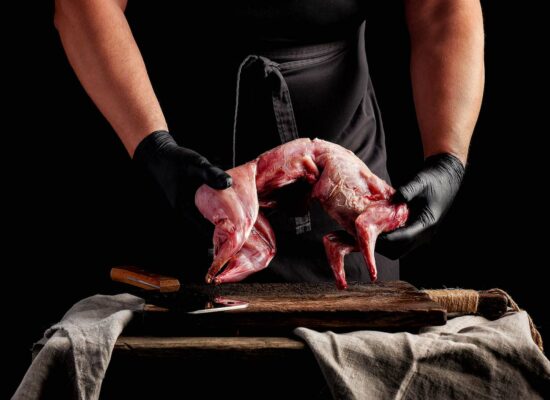 A person wearing black gloves and a dark apron holds a skinned rabbit over a wooden cutting board on a rustic table. A knife with a wooden handle rests to the side, and a cloth is draped over the table. The dark background emphasizes the scene, reminiscent of an old Iowa kitchen stocked by WG-Provisions.