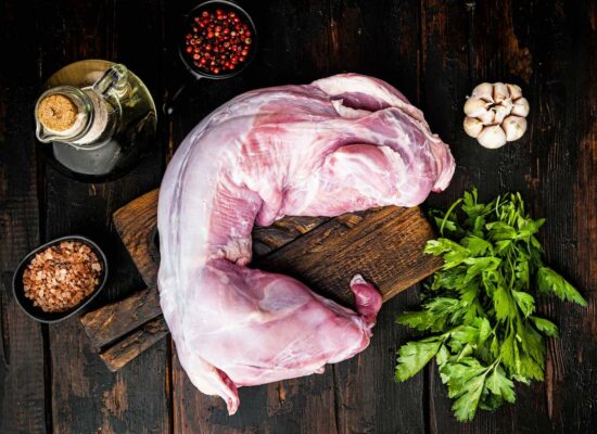 A raw, skinned rabbit lies on a wooden cutting board surrounded by cooking ingredients from WG-Provisions in Iowa. Nearby are pink Himalayan salt, red peppercorns, a bottle of oil, a bulb of garlic, and a bunch of fresh parsley on a dark wooden surface.