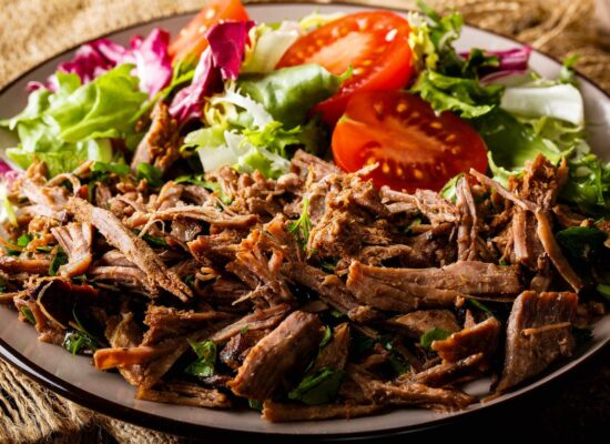 A plate of shredded beef served with fresh mixed greens and slices of ripe tomatoes. The colorful salad, featuring green and purple lettuce varieties, is presented on a rustic, woven surface. This WG-Provisions specialty highlights the flavors of Iowa farm-fresh produce.
