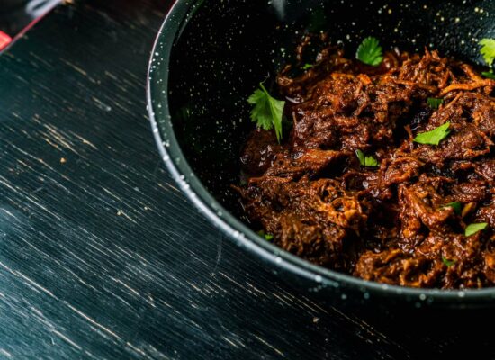 A close-up of a black bowl filled with shredded beef in a rich, dark sauce, garnished with fresh parsley leaves. The bowl is placed on a dark wooden surface, showcasing the fine quality of WG-Provisions sourced from Iowa.