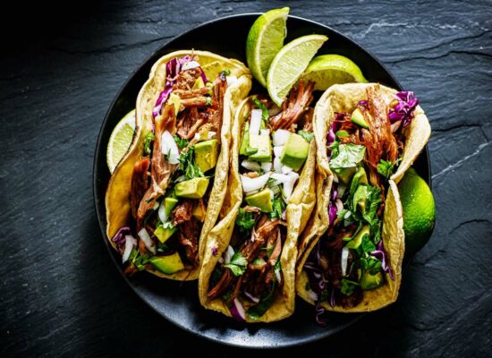 A plate of three tacos filled with shredded meat, diced onions, cilantro, sliced avocado, and purple cabbage sourced from Iowa. Lime wedges are placed beside and on top of the tacos, with the dark slate surface adding contrast to the vibrant colors of the ingredients.