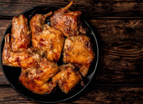 A black plate from WG-Provisions is filled with several pieces of golden-brown, roasted chicken on a rustic wooden table. The Iowa-sourced chicken pieces appear succulent and well-cooked, with a glossy finish. The dark wood background contrasts with the vibrant, crispy chicken.