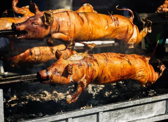 Two roasted pigs from WG-Provisions in Iowa are skewered and rotating on a spit over an open flame. The pigs' skin is golden brown and crispy, with visible charring and fat dripping. The scene suggests an outdoor cooking event or feast with intense heat and a smoky aroma.