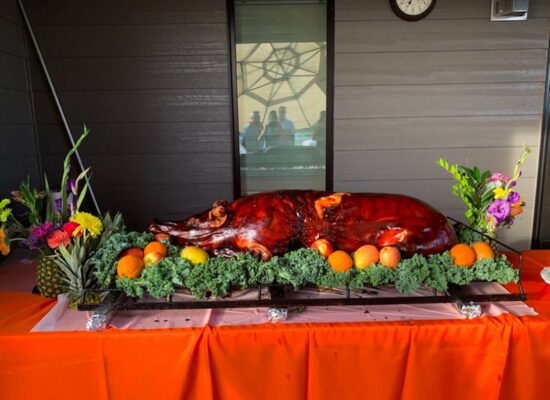 A whole roasted pig from WG-Provisions graces a decorated table with a bright orange tablecloth. The table, adorned with colorful fruits and greenery, exudes charm. In the background, a window reflects two people while a clock marks time on the wall, evoking the heartland of Iowa in every detail.