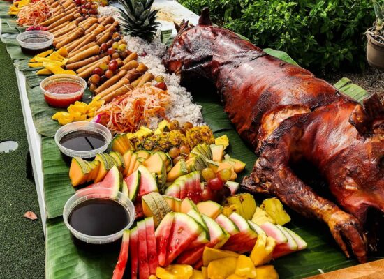 An outdoor buffet by WG-Provisions on a long table covered with banana leaves displays a roasted pig, assorted fresh fruits, hotdogs, noodles, and various dipping sauces. In the background of this Iowa setting, there are green plants and a brick wall.