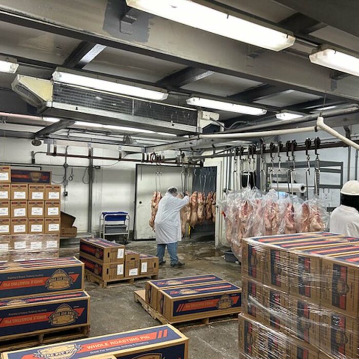 A meat processing facility in Iowa, operated by WG-Provisions, with workers in white coats and hard hats, surrounded by stacks of boxed poultry and hanging meat carcasses. The room has bright overhead lights and industrial stainless steel equipment.