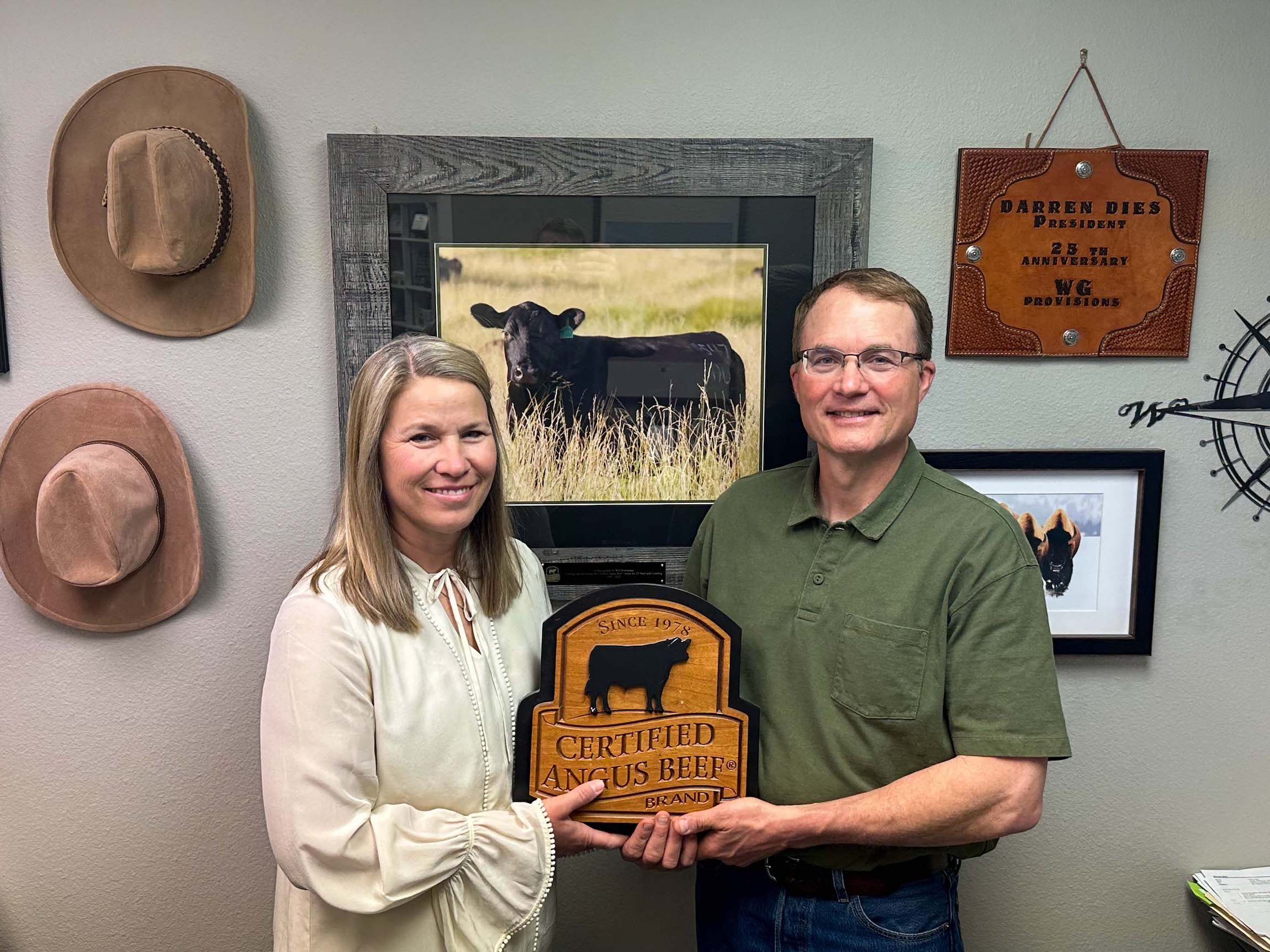 A woman and a man, both smiling, hold a "Certified Angus Beef" plaque. Behind them is a framed picture of a black Angus cow. They are standing in a room with hats on the wall and a name plaque for "Darren Dies.
