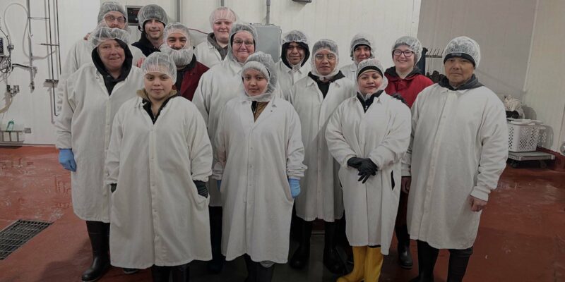 A group of 14 people, wearing white lab coats, hairnets, and various colored boots, stand together in what appears to be a cleanroom or laboratory environment at WG-Provisions in Iowa. The floor is red and the walls are white. They all look at the camera, some smiling.